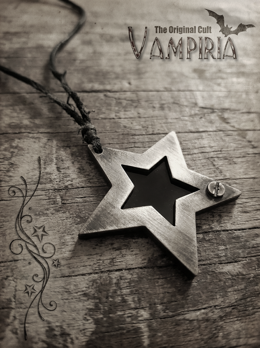 Sternförmiger Metallanhänger mit schwarzem Einsatz an schwarzem Band auf Holzoberfläche, oben rechts Vampiria-Logo, links dekoratives Ornament.