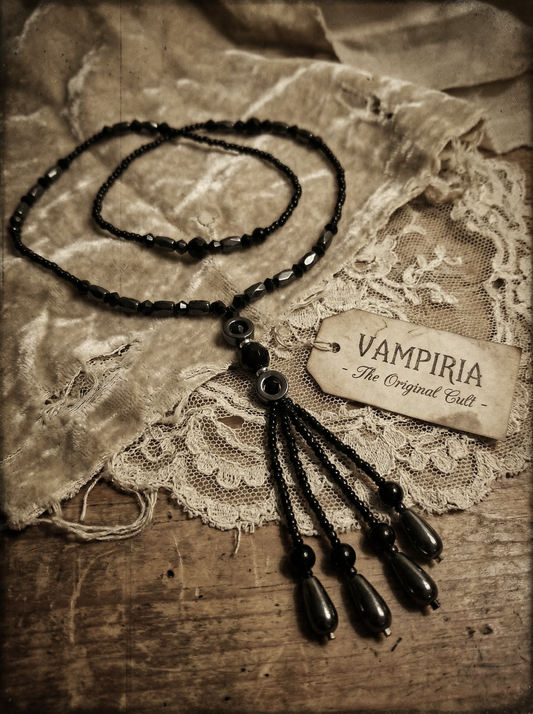Schwarzes Y-Collier mit tropfenförmigen Hämatit-Perlen und kleinen schwarzen Perlen auf heller Spitze und Stoff, daneben Etikett mit „Vampiria“.