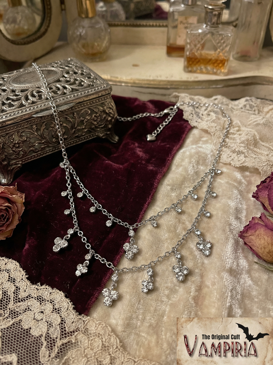 Silberfarbenes filigranes Collier mit mehreren Kettenreihen und kleinen klaren Crystal-Anhängern auf cremefarbenem und bordeauxfarbenem Stoff neben einer verzierten Schmuckdose.
