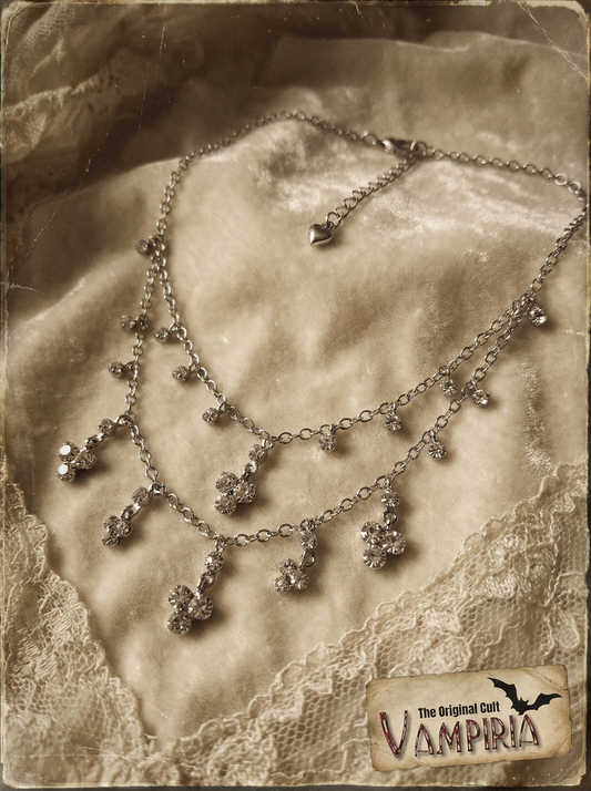 Mehrreihiges silberfarbenes Collier mit Crystal-Anhängern auf hellem Samtstoff im Vintage-Stil, mit Spitzenstoff und Vampiria-Logo im Bild.
