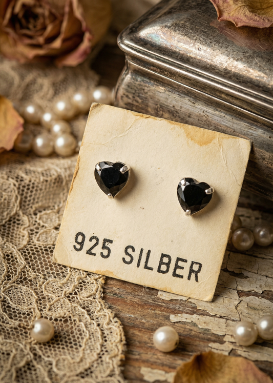 Schwarze Herz-Ohrstecker aus 925 Silber auf Schmuckkarte neben Vintage-Dekoration.