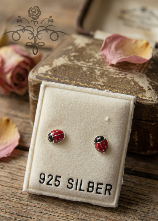 Zwei Mini Ohrstecker in Marienkäferform aus 925 Silber mit roten und schwarzen Details auf einer Schmuckkarte mit Aufdruck „925 SILBER“, umgeben von Rosen und Vintage-Flakons.