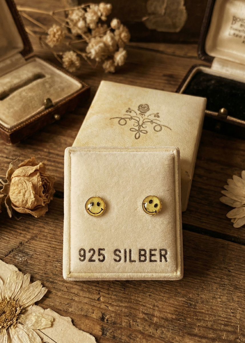 Mini Ohrstecker mit gelbem Smilie-Motiv in 3D-Optik auf hellem Schmuckdisplay vor Vintage-Dekoration aus Holz, Schmuckschachteln und Trockenblumen.