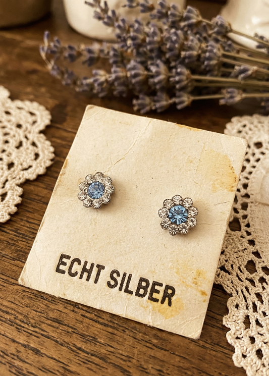 Zwei kleine Blumen-Ohrstecker mit hellblauem Stein in der Mitte auf einer Karte mit Aufschrift „ECHT SILBER“, daneben Lavendel und Häkeldeckchen auf Holztisch.