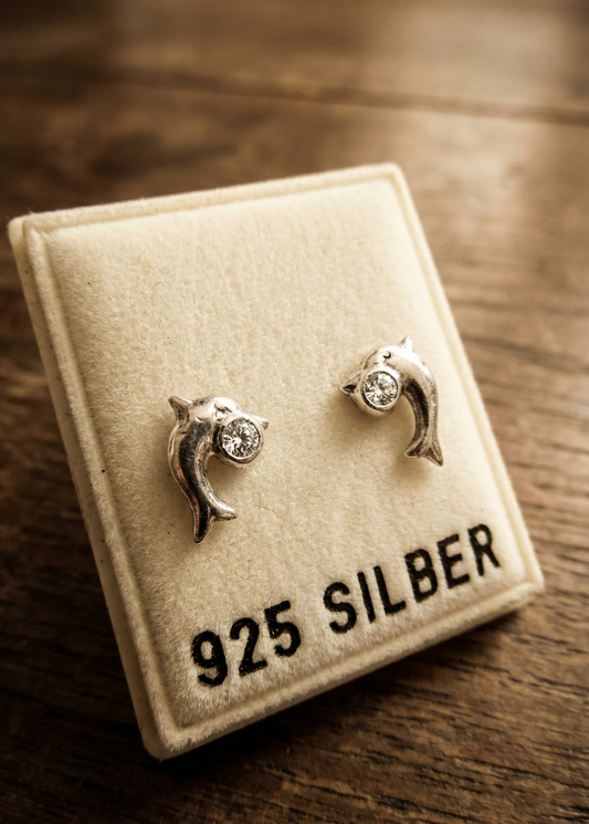 Zwei kleine silberfarbene Delfin-Ohrstecker mit klarem Kristall auf einer beigen Schmuckkarte mit Aufschrift „925 SILBER“ auf Holzuntergrund.