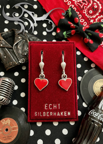 Zwei rote Herz-Ohrringe mit silberfarbenen Bügeln auf rotem Samt-Display mit Aufschrift „ECHT SILBERHAKEN“, umgeben von Schallplatten, Mikrofon und Rockabilly-Accessoires.