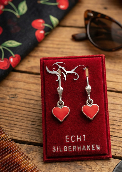 Zwei rote Herz-Ohrringe mit silberfarbenen Bügeln auf rotem Samt-Display mit Aufschrift „ECHT SILBERHAKEN“ auf Holztisch, daneben Sonnenbrille und gemusterter Stoff.