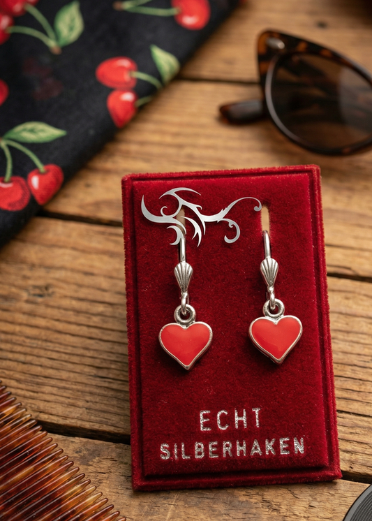 Zwei rote Herz-Ohrringe mit silberfarbenen Bügeln auf rotem Samt-Display mit Aufschrift „ECHT SILBERHAKEN“ auf Holztisch, daneben Sonnenbrille und gemusterter Stoff.