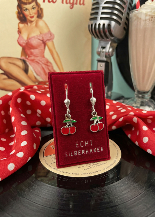 Rote Kirsch-Ohrringe auf rotem Samtkärtchen mit Aufschrift „ECHT SILBERHAKEN“, dekoriert mit Schallplatten, Mikrofon, schwarzem Lederjäckchen, rotem Tuch mit Punkten und Coca-Cola-Flasche.