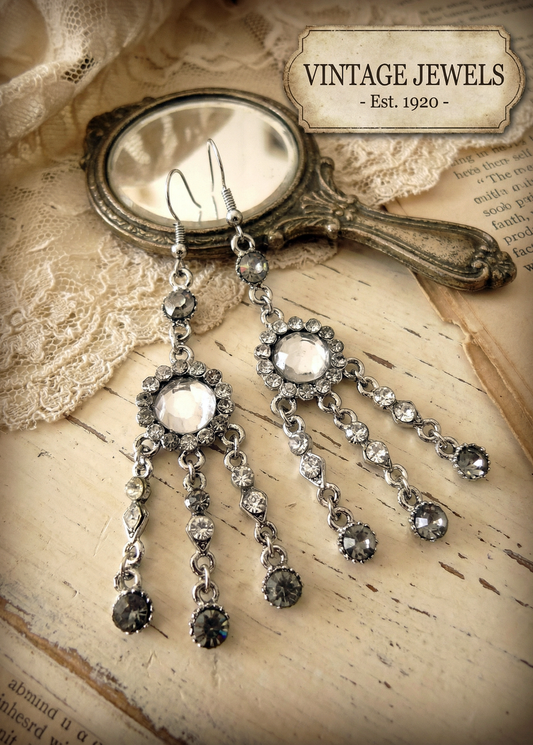 Zwei lange silberfarbene Vintage-Ohrhänger mit klaren und dunklen Strasssteinen liegen auf einer hellen Holzfläche neben einem verzierten Handspiegel und Spitze; oben rechts ein Schild mit der Aufschrift „VINTAGE JEWELS - Est. 1920 -“.
