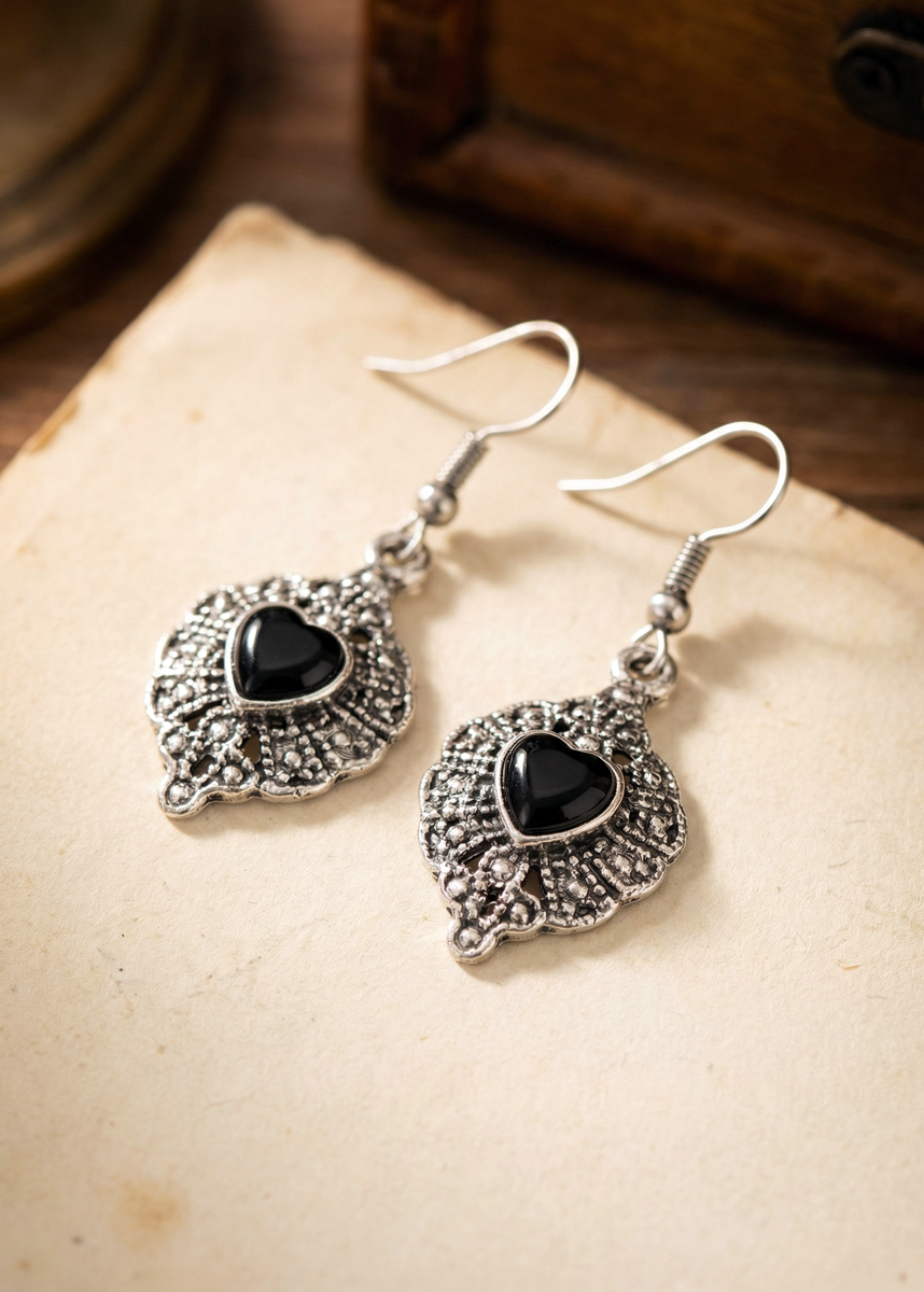 Zwei silberfarbene Hängeohrringe mit ornamentaler barocker Form und schwarzem Herz in der Mitte auf hellem Untergrund.