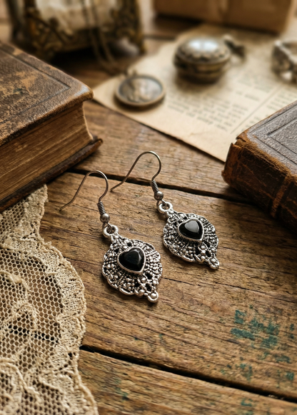 Zwei silberfarbene barocke Herz-Ohrringe mit schwarzer Herzfüllung auf rustikalem Holztisch zwischen Büchern und Spitze.