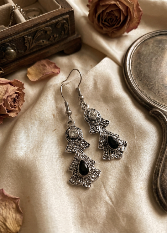 Silberfarbene barocke Hängeohrringe mit schwarzen tropfenförmigen Steinen und klaren Strasssteinen auf cremefarbenem Stoff neben Spiegel und getrockneten Rosen.