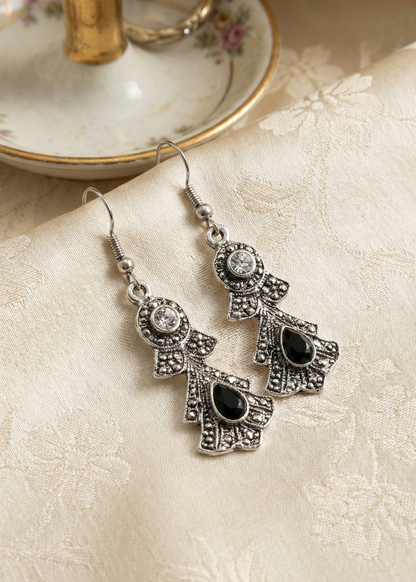 Zwei silberfarbene barocke Hängeohrringe mit schwarzen Tropfensteinen und klaren Strasssteinen auf hellem gemustertem Stoff neben Porzellanständer.