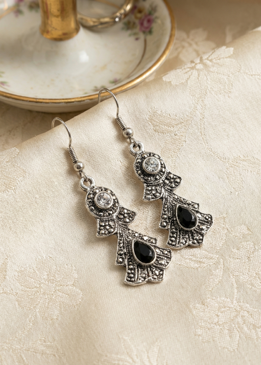 Zwei silberfarbene barocke Hängeohrringe mit schwarzen Tropfensteinen und klaren Strasssteinen auf hellem gemustertem Stoff neben Porzellanständer.