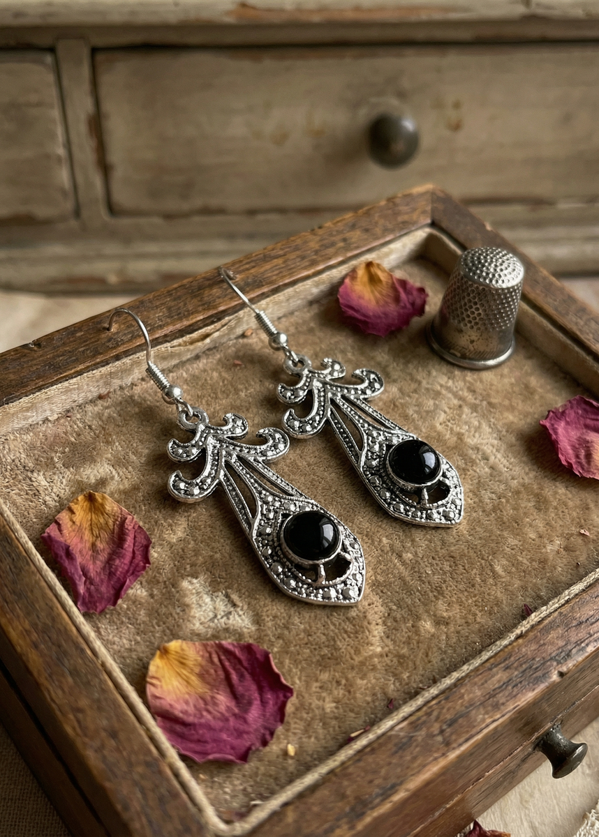 Zwei barocke Hängeohrringe in antik-silberfarbener Optik mit schwarzem Cabochon liegen in einer alten Holzschublade auf Samt, daneben Rosenblätter und ein Fingerhut.