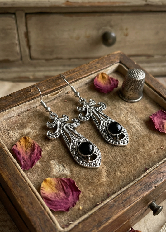 Zwei barocke Hängeohrringe in antik-silberfarbener Optik mit schwarzem Cabochon liegen in einer alten Holzschublade auf Samt, daneben Rosenblätter und ein Fingerhut.