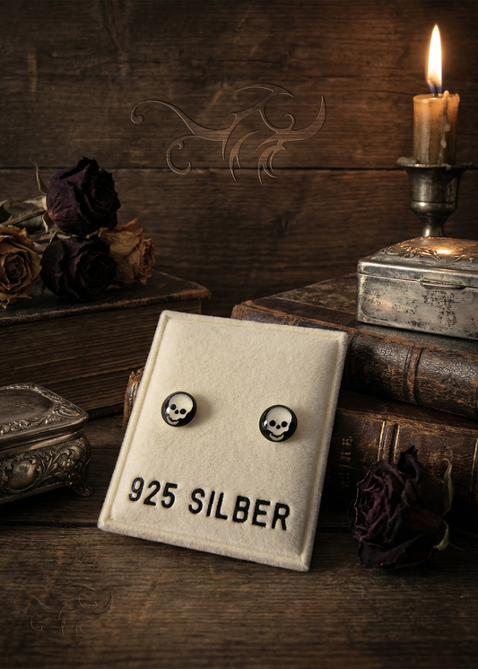 Zwei runde Mini-Ohrstecker mit schwarz-weißem Totenkopfmotiv auf einer cremefarbenen Schmuckkarte mit Aufschrift „925 SILBER“, arrangiert vor dunklem Holz, Kerze, alten Büchern und getrockneten Rosen.