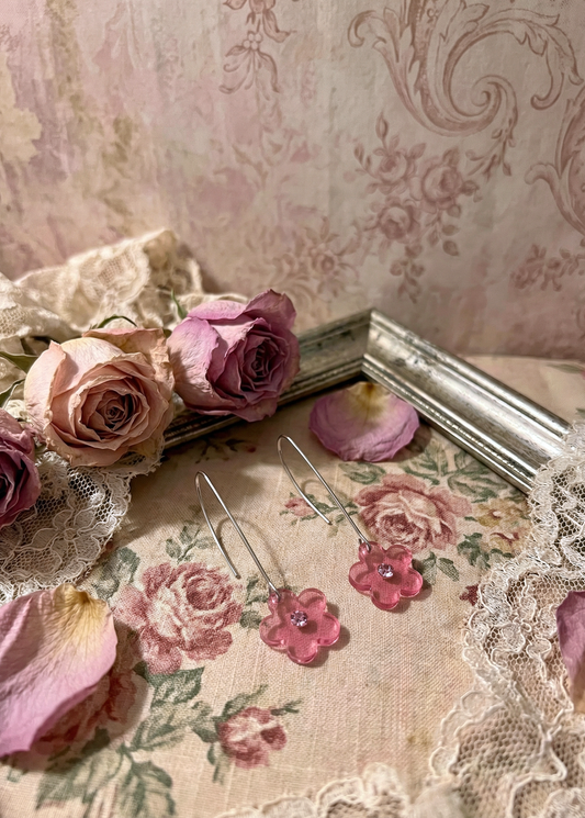 Zwei rosa Blüten-Hängeohrringe mit silberfarbenen Haken auf floralem Stoff mit Rosen, Spitze und Bilderrahmen.