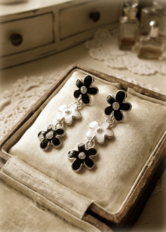 Schwarz-weiße Vintage-Blumenohrringe mit silberfarbenem Rand und kleinen klaren Steinen, präsentiert in einer geöffneten Schmuckschatulle auf hellem Stoff.