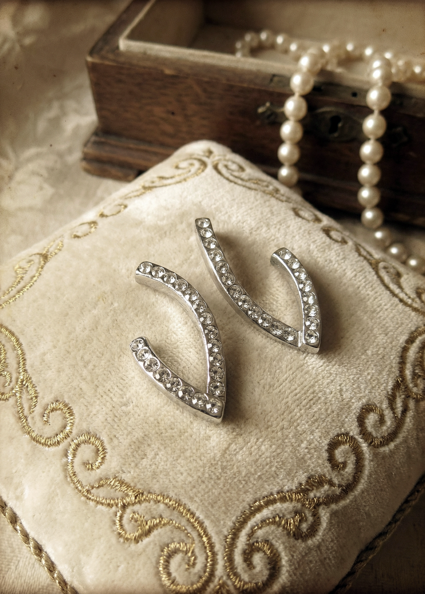 Zwei silberfarbene Design-Ohrstecker in geschwungener V-Form mit klaren Strasssteinen auf einem bestickten cremefarbenen Schmuckkissen, Perlenkette und Holzschatulle im Hintergrund.