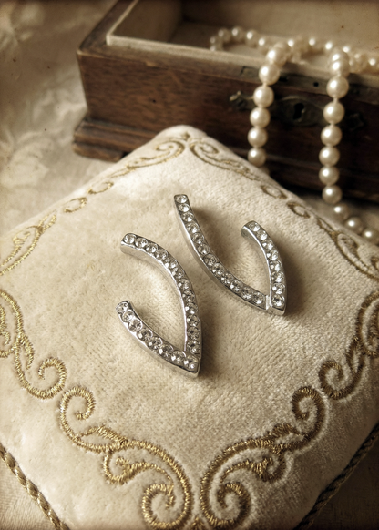 Zwei silberfarbene Design-Ohrstecker in geschwungener V-Form mit klaren Strasssteinen auf einem bestickten cremefarbenen Schmuckkissen, Perlenkette und Holzschatulle im Hintergrund.