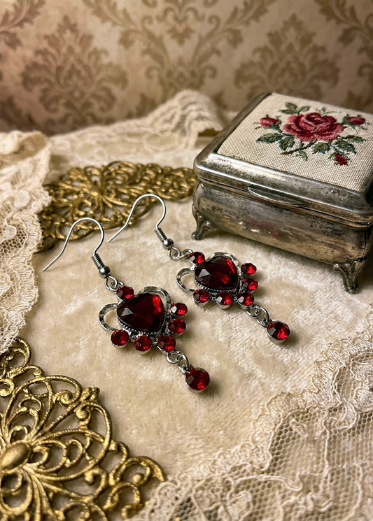 Rote Herz-Ohrringe mit roten Strasssteinen und kleinem hängenden roten Stein auf heller Samtfläche neben Spitze und Vintage-Schmuckdose.