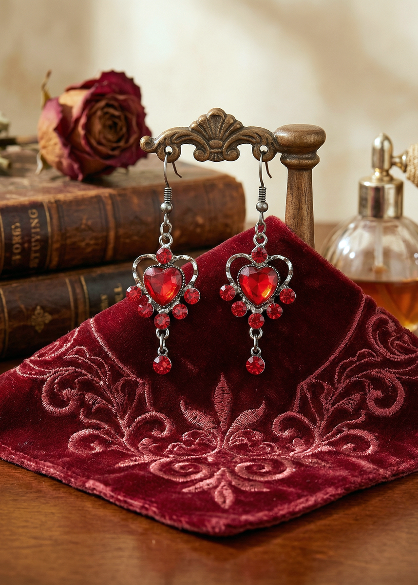 Rote Herz-Ohrringe mit Herzstein und roten Strassdetails hängend an kleinem Holzständer vor Büchern und Parfumflakon.