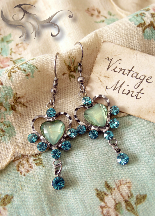 Zwei silberfarbene Vintage-Ohrringe mit mintgrünem Herzstein, türkisblauen Kristallen und kleinem hängenden Kristall auf gemustertem Stoff neben Karte mit Aufschrift „Vintage Mint“.