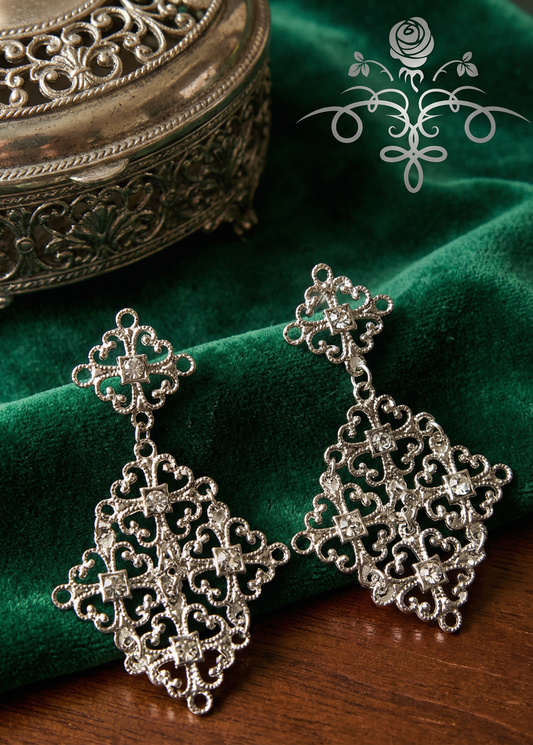 Zwei silberfarbene barocke Ornament-Ohrringe mit Glitzerdetails auf grünem Samt vor dekorativer Schmuckdose.