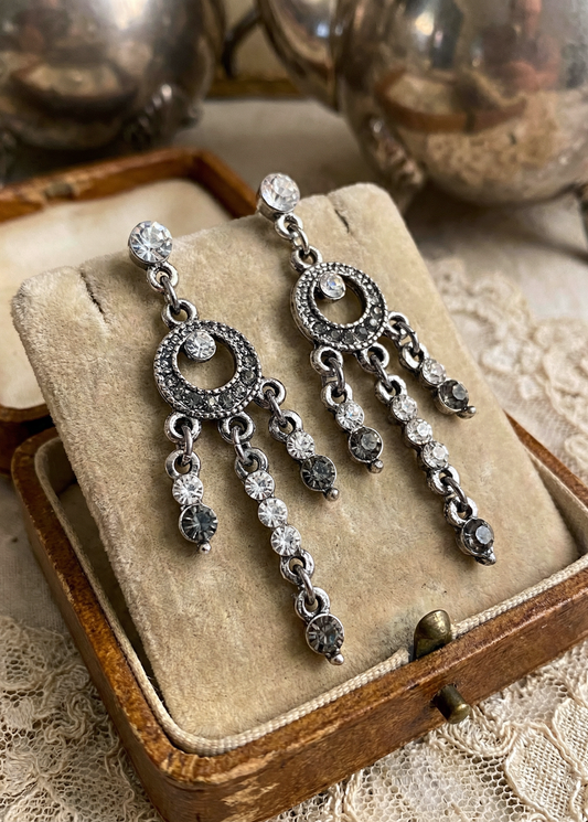 Zwei silberfarbene Hängeohrringe im Boho-Vintage-Stil mit runden Strass-Elementen und mehreren beweglichen Strass-Ketten auf einer beigen Schmuckschatulle.