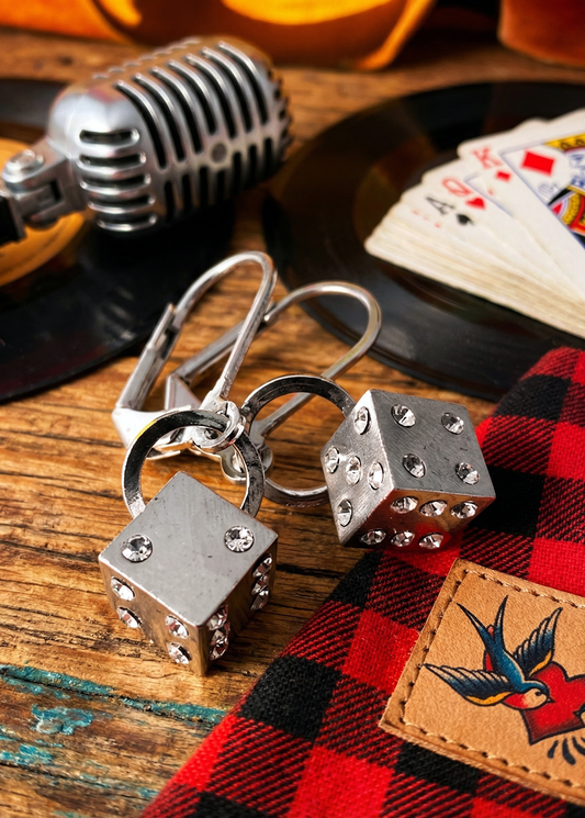 Zwei silberfarbene Würfel-Ohrringe mit Strasssteinen und Klappverschluss liegen auf einem Holztisch neben Mikrofon, Schallplatte und Spielkarten.