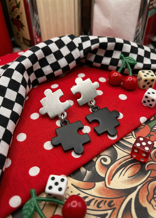 Zweifarbige Puzzle-Ohrstecker mit gebürstetem silberfarbenem Stecker und schwarzem Puzzle-Anhänger auf rotem gepunktetem Tuch mit Schachbrettmuster, Würfeln und Kirschen.